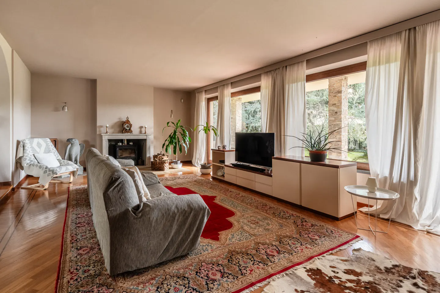 Living room with hardwood floors, a gray sofa, and a large patterned rug. A fireplace, TV, and windows with white curtains are also visible.