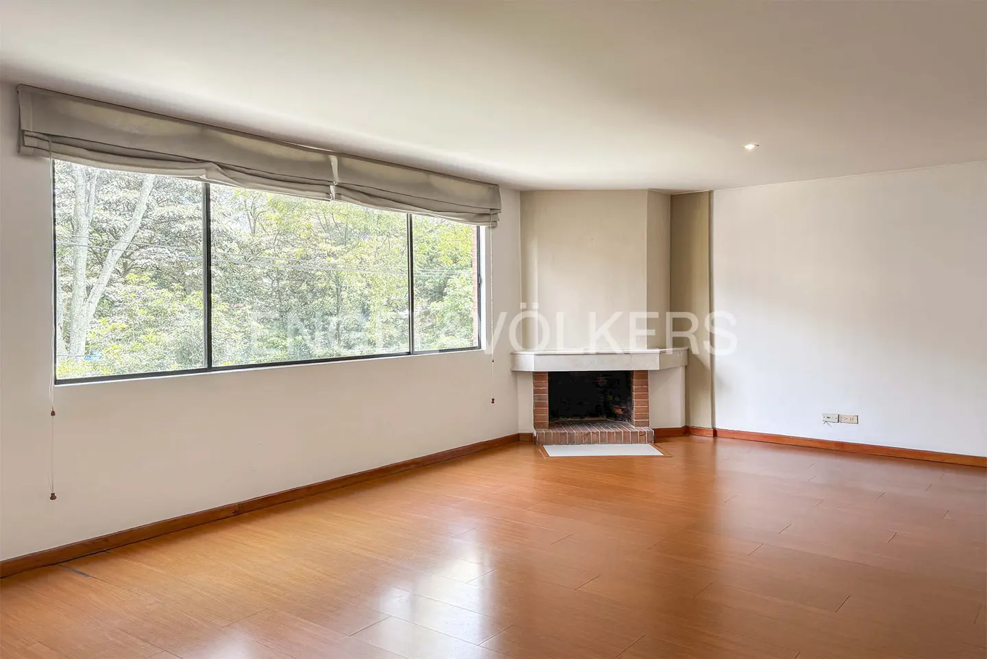 Bright living room with hardwood floors, a large window with a view of trees, and a brick fireplace.