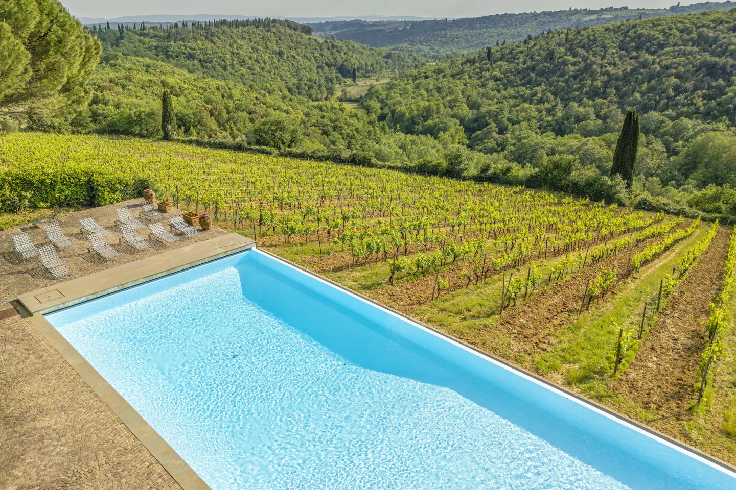 A bright blue infinity pool overlooks a lush green vineyard and rolling hills in the Italian countryside.