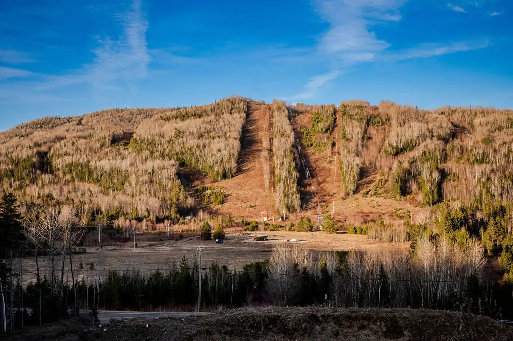 Ski hill with bare runs cutting through a forest on a sunny day. Blue sky with wispy clouds above. Base area visible at the bottom of the hill.