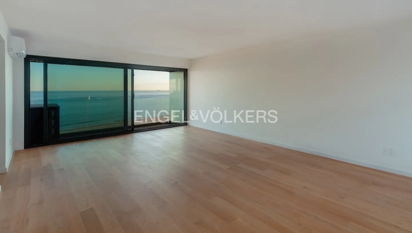 Empty room with light wood floors, white walls, and a large sliding glass door with ocean view. Engel & Völkers logo in the center.