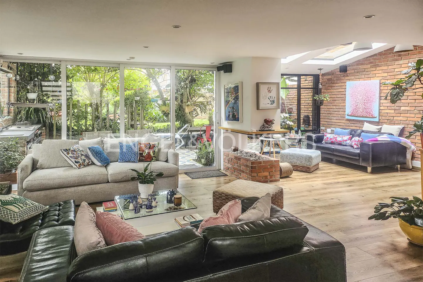 Bright living room with hardwood floors, sofas, and a glass wall overlooking a lush garden. Artwork adorns the walls.