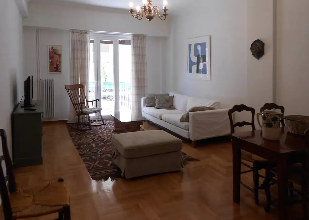 Bright living room with hardwood floors, white sofa, rocking chair, and dining table. A patterned rug adds warmth. Balcony doors let in natural light.