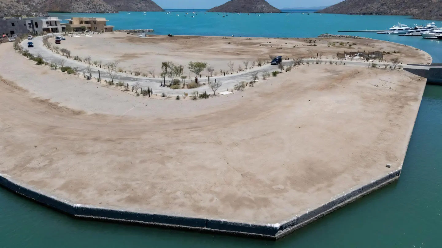 Aerial view of a sandy peninsula with a road, buildings, and turquoise water. Mountains are visible in the background.