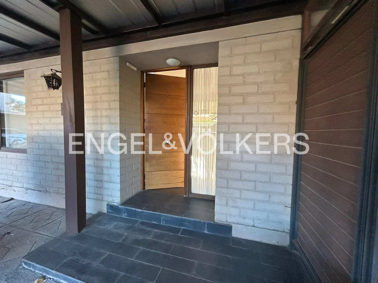 Exterior view of a brick house with an open wooden front door and a black tiled porch. The Engel & Volkers logo is superimposed on the image.