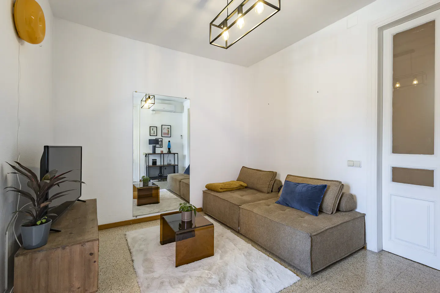 Modern living room with a tan modular sofa, blue pillow, glass coffee table, white rug, TV, and a large wall mirror.