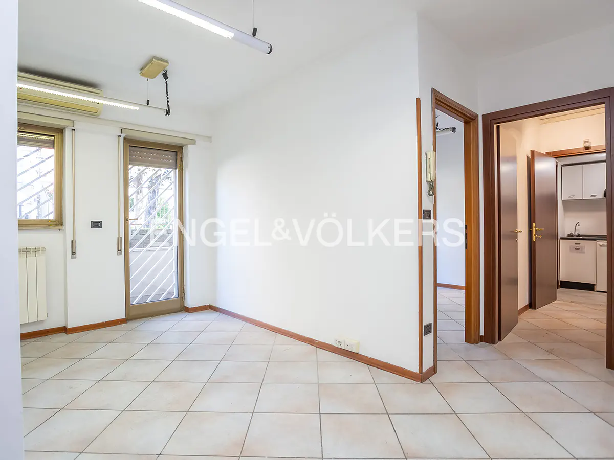 Bright, empty room with tile floor, white walls, and wood trim. A window and door lead outside. A doorway leads to a kitchen.