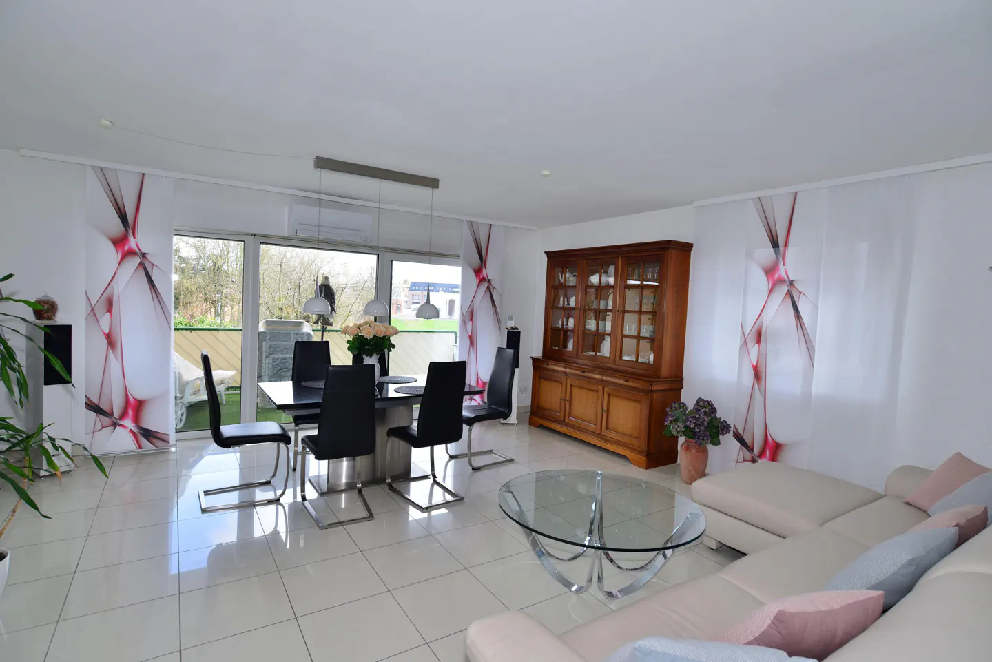 Bright living room with white tile floors, a black dining table with chairs, and a light-colored sectional sofa. A wooden hutch stands against the wall.