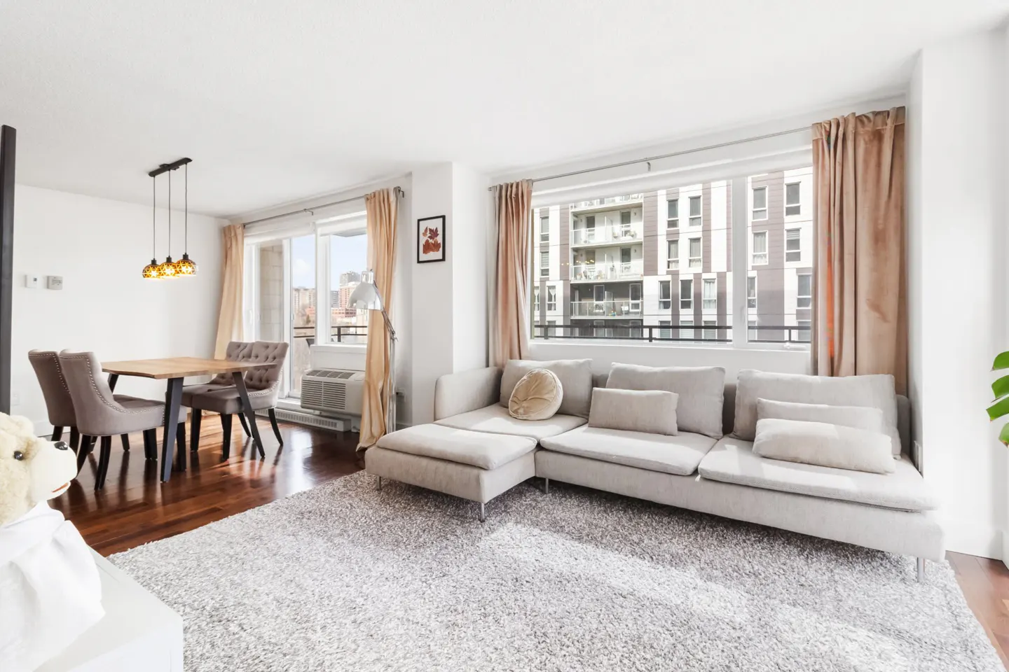 Bright living room with a beige sectional sofa, dining table with chairs, and a large window with peach curtains.