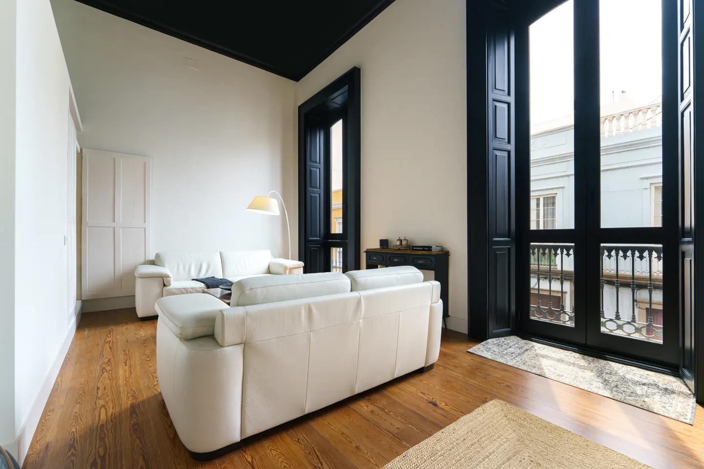 Bright living room with white walls, dark trim, and wood floors. White sofas face large black-framed windows with a view of a building exterior.