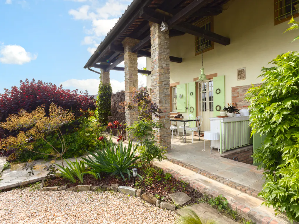 Exterior view of a cream-colored house with green shutters and stone columns. A patio with a table and chairs is visible. The garden is lush with colorful plants.
