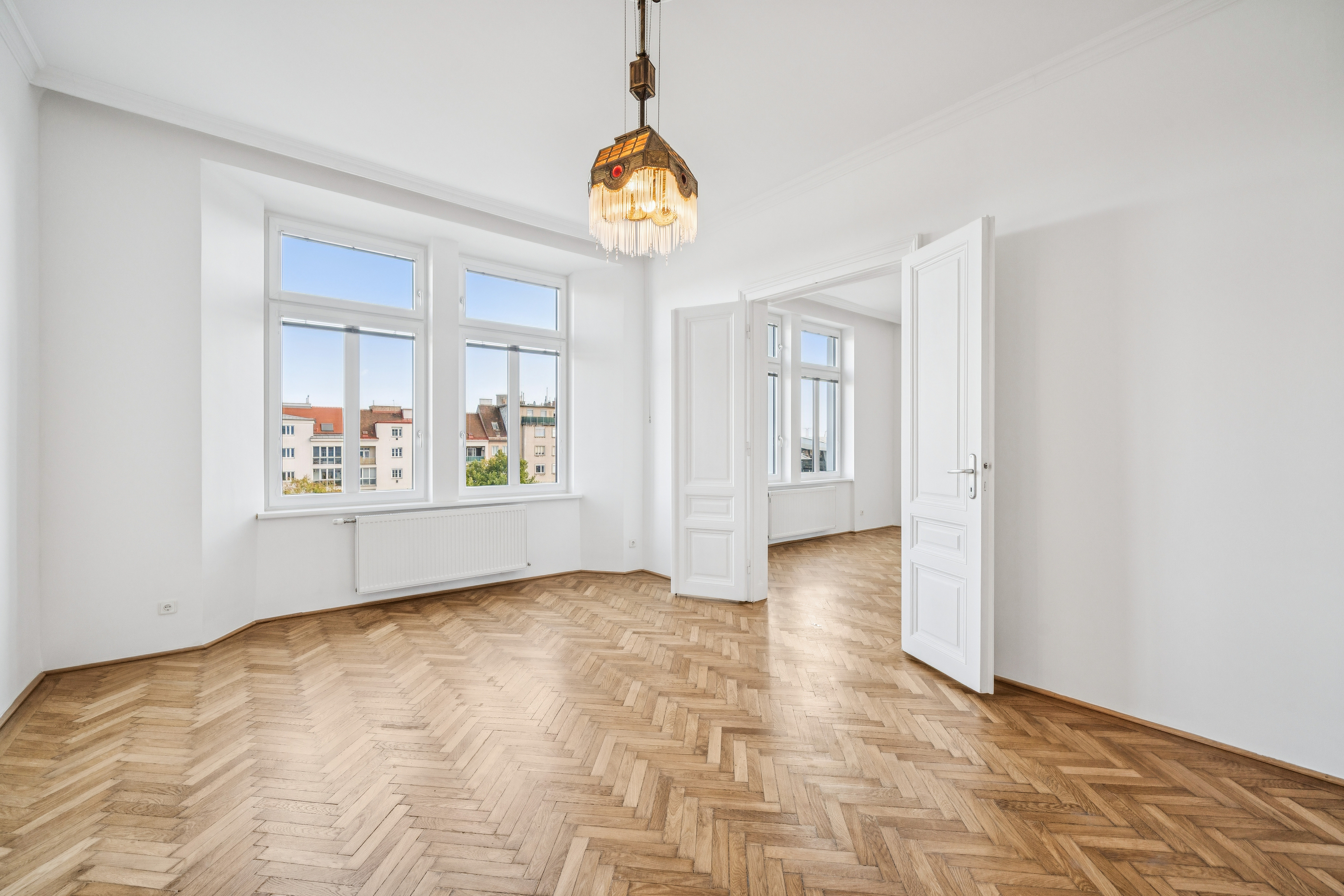 Empty room with white walls, herringbone wood floors, large windows, and a vintage-style chandelier. [AI-generated text]