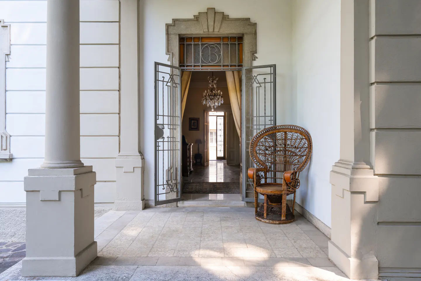 Covered porch with white columns, stone floor, and open metal gate leading to a chandelier-lit interior. A wicker peacock chair sits to the right.
