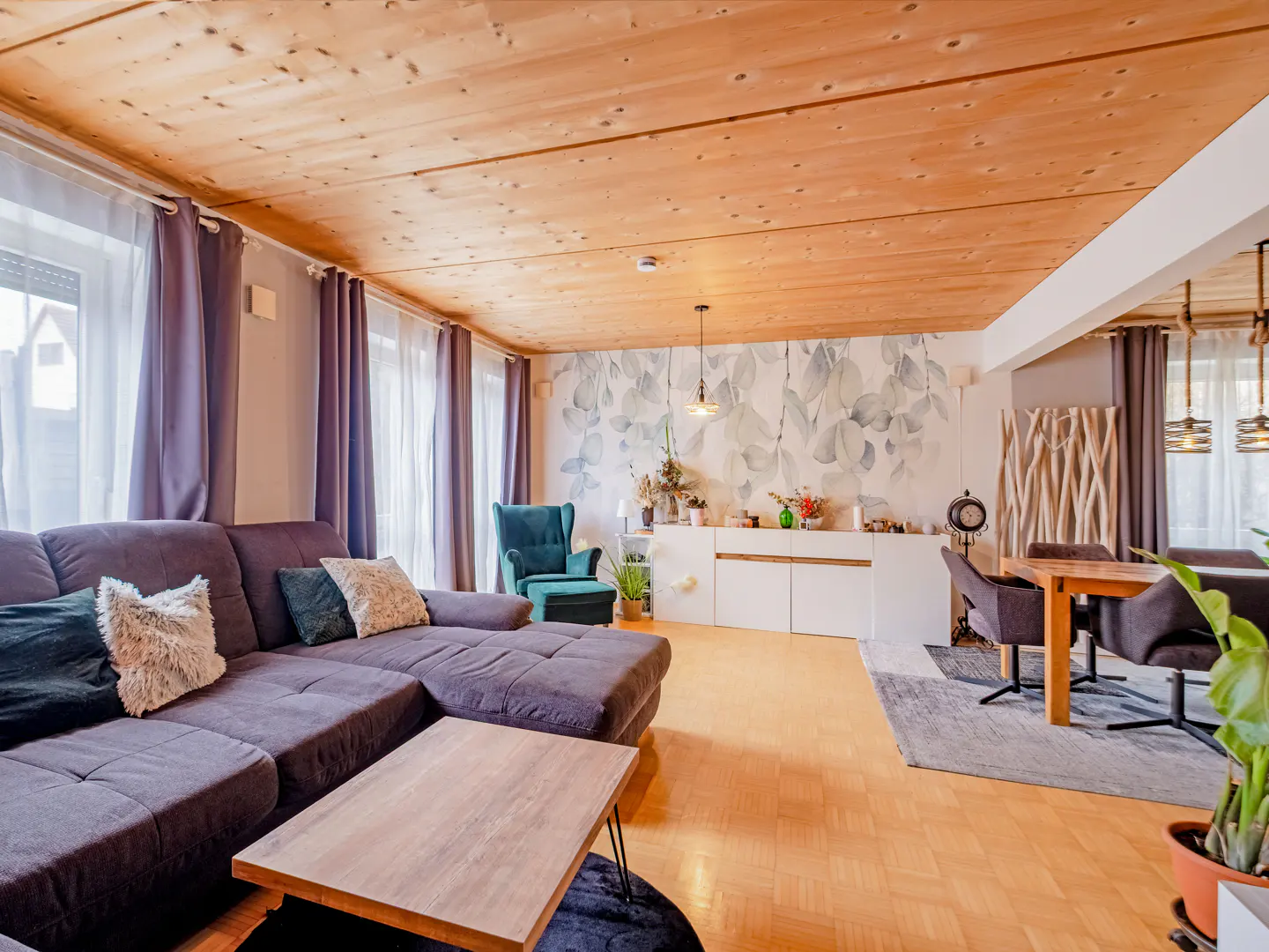 Living room with wood ceiling, purple sofa, and floral wallpaper. A teal armchair and dining table are also visible.