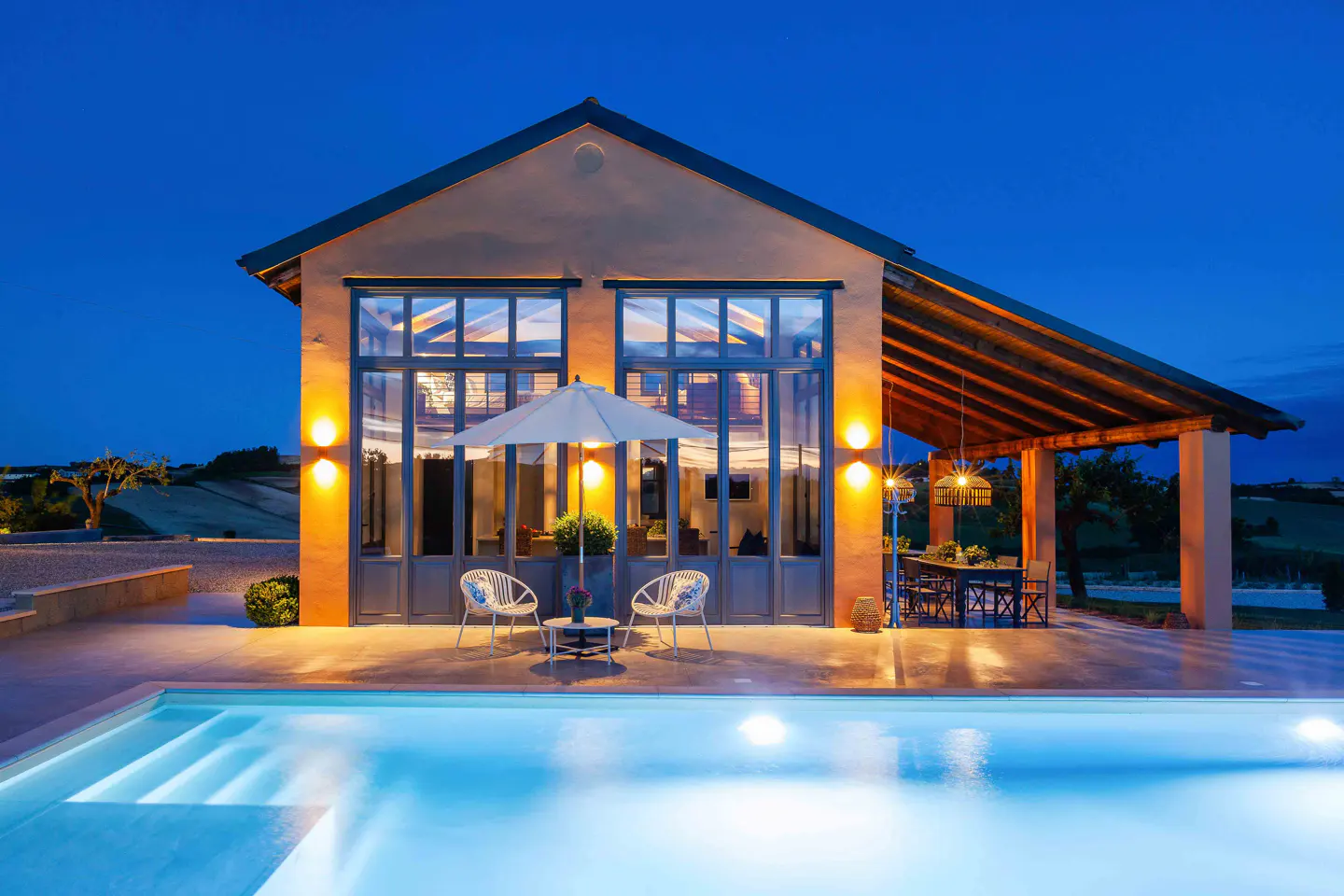 Exterior view of a tan house with large windows, a pool, and outdoor seating at dusk.