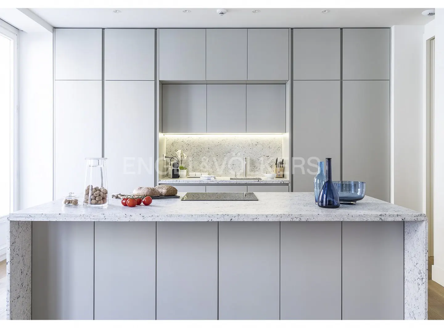 A modern kitchen with gray cabinets, a speckled countertop island with bread, tomatoes, and blue glass vases.