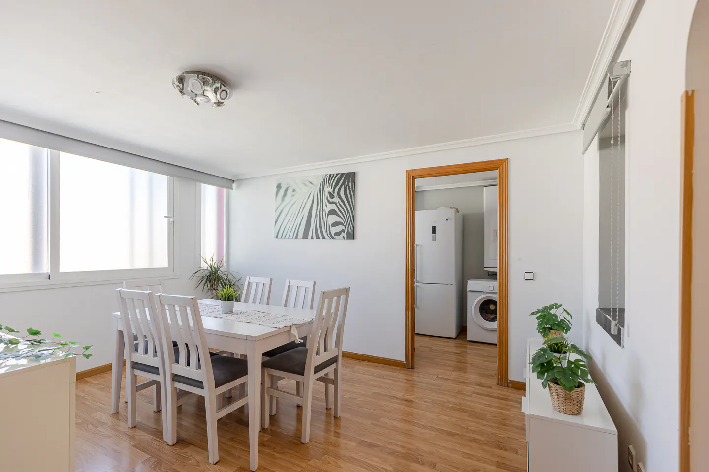 Bright dining room with white table, chairs, and walls. A zebra print hangs on the wall. A doorway leads to a laundry area with a washer and refrigerator.
