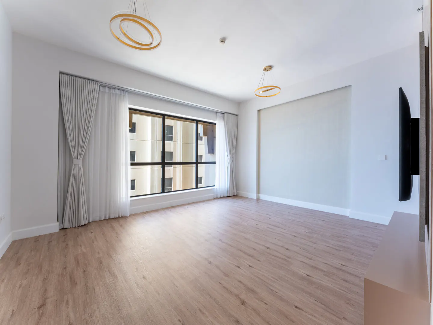 Bright, empty living room with wood floors, white walls, and a large window with white curtains. Two modern, circular chandeliers hang from the ceiling.