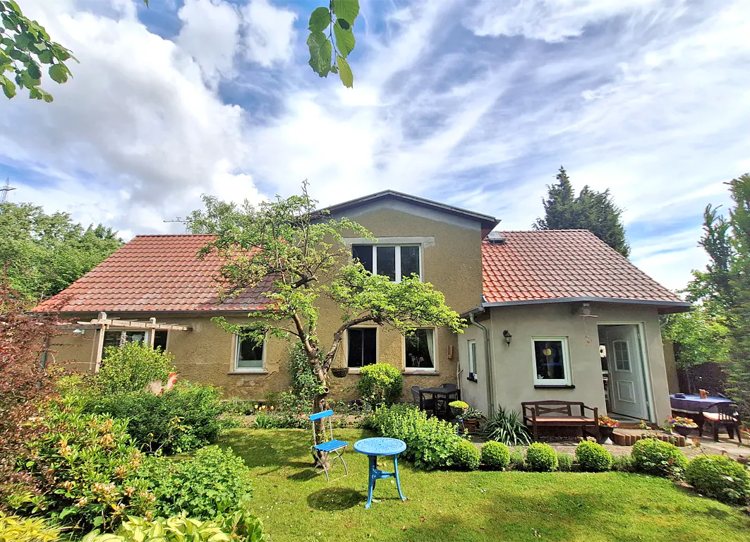 Exterior view of a tan house with a red tile roof, green lawn, and blue outdoor table and chairs.