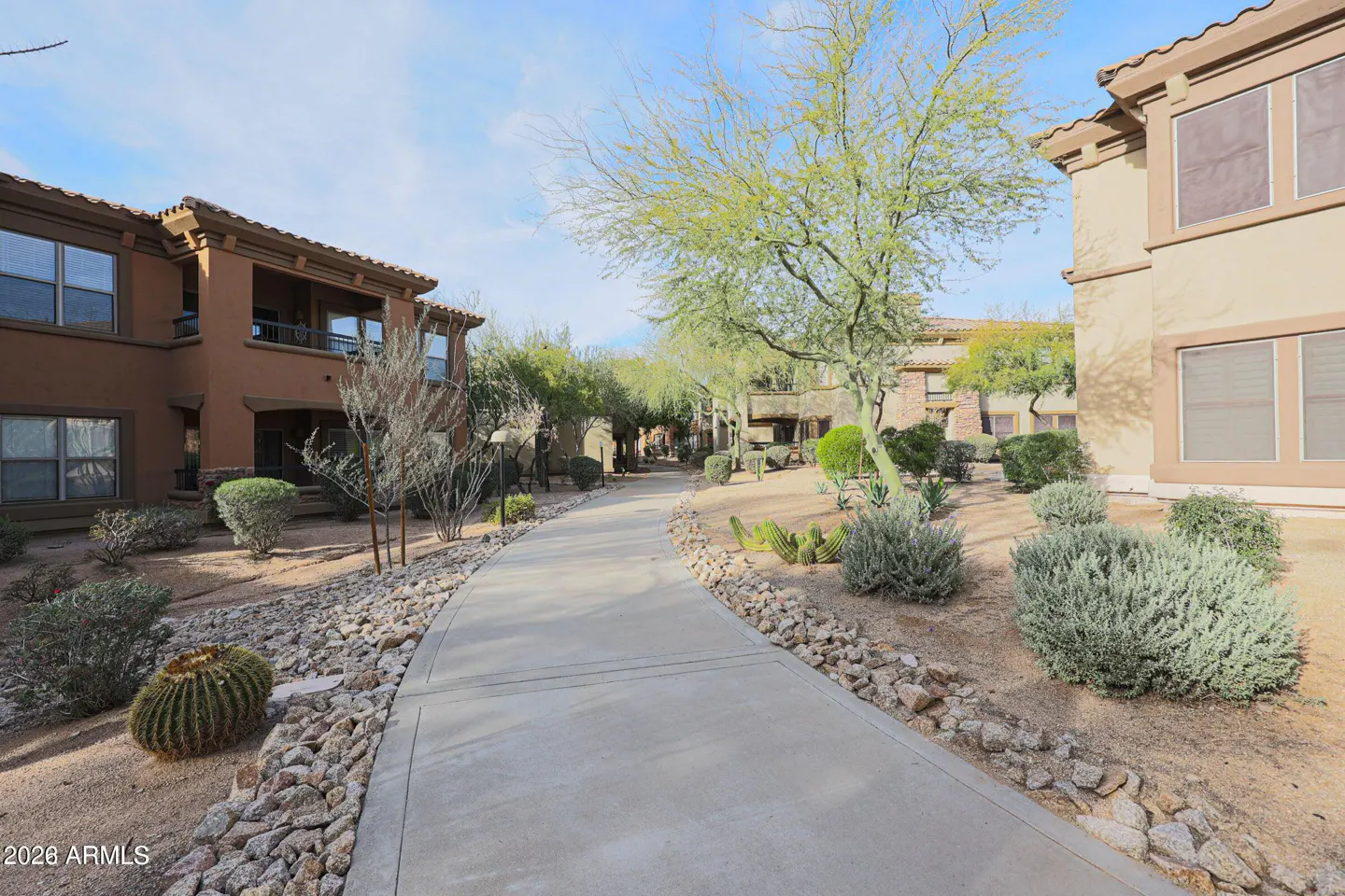 Exterior view of a desert-landscaped apartment complex with a concrete walkway, cacti, and desert plants. The buildings are tan and brown.