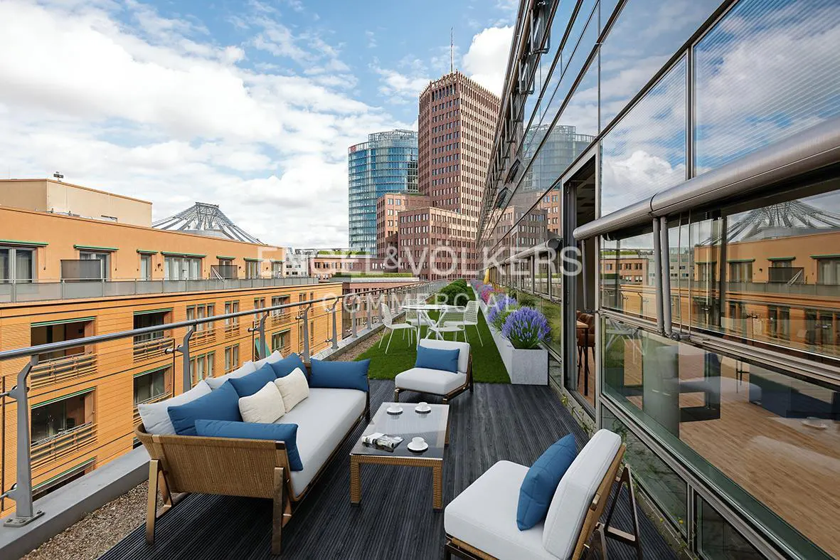Rooftop patio with a sofa, chairs, and a table. City buildings are visible in the background. Blue and white cushions add color.
