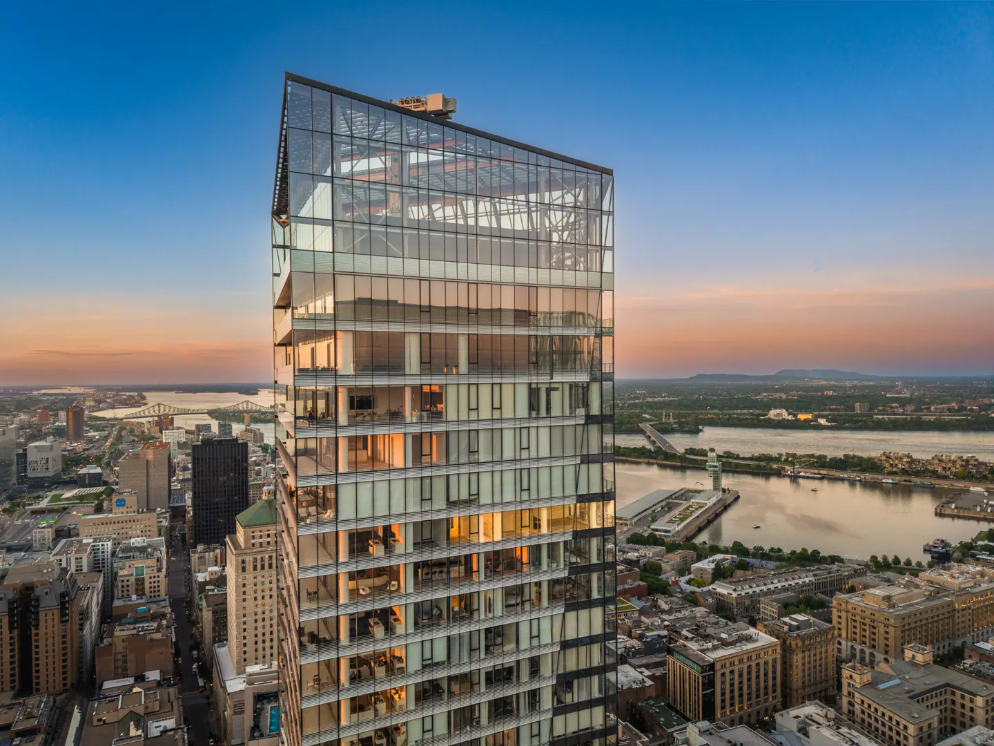 Modern glass skyscraper at sunset overlooking a city skyline and river, with warm interior lights visible in the units.