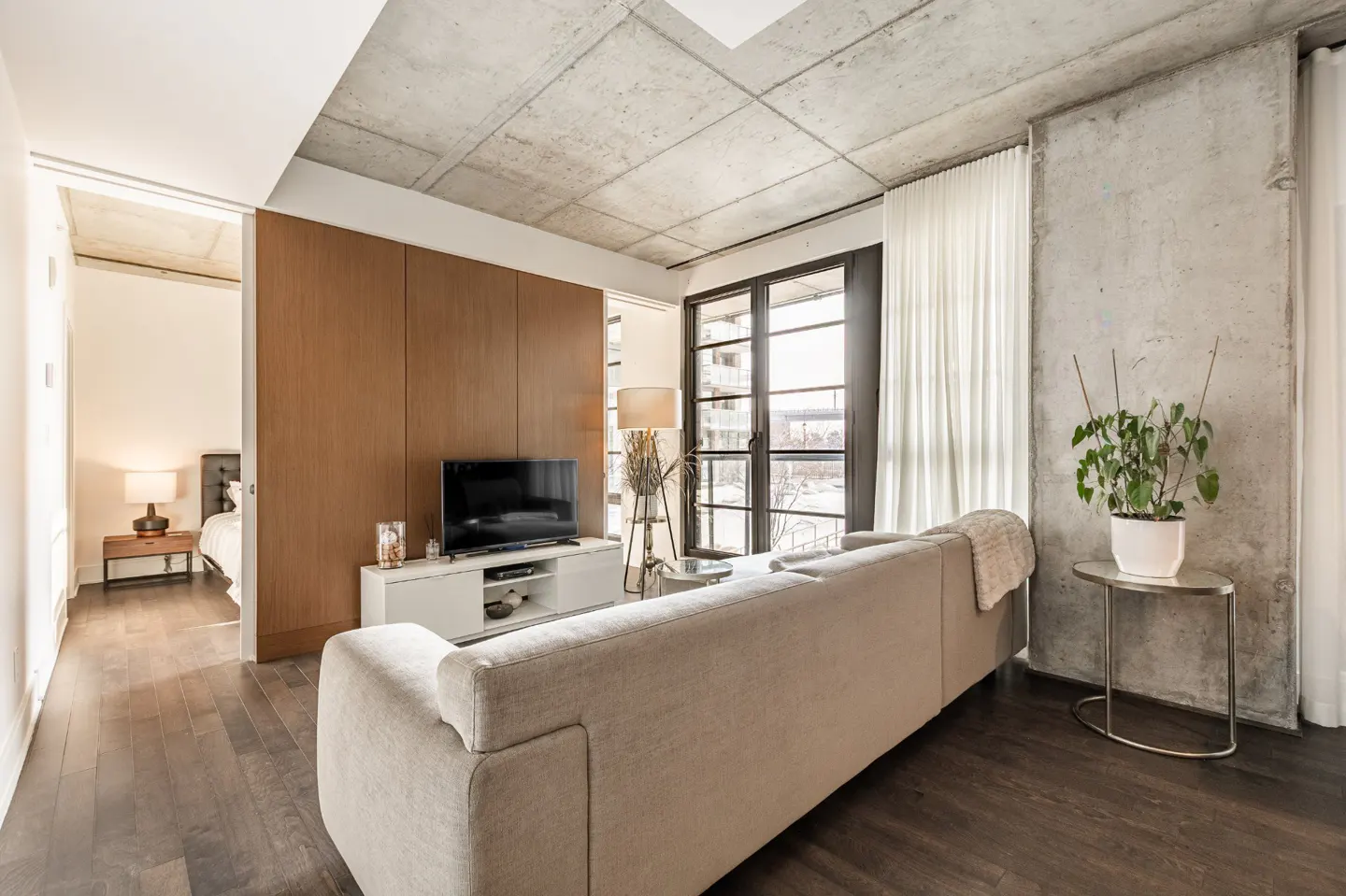 Living room with concrete ceiling and walls, dark wood floors, beige sofa, TV, and a view from the window.
