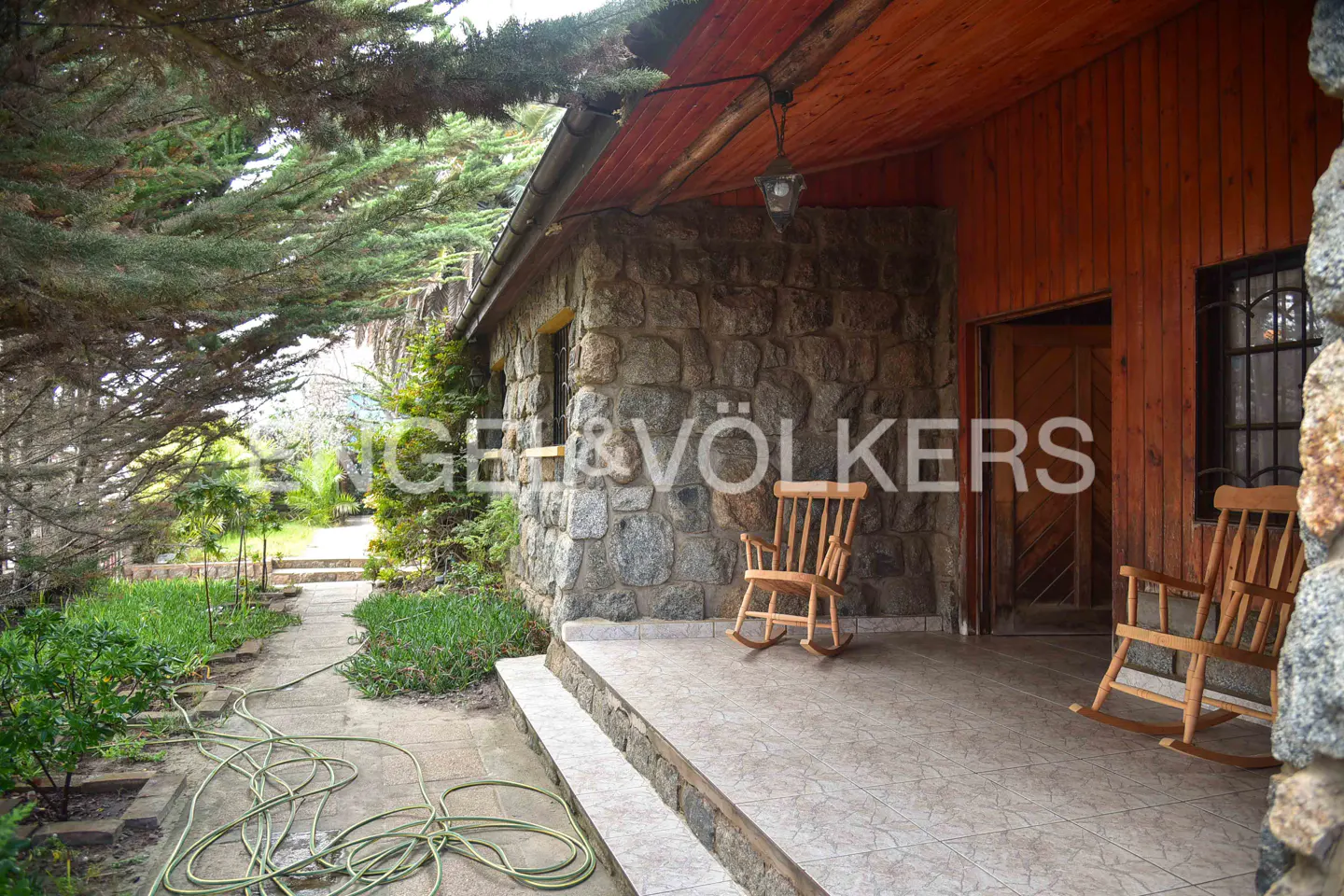 Stone house with a wood porch and two rocking chairs. A garden path leads to the house. The Engel & Völkers logo is visible.