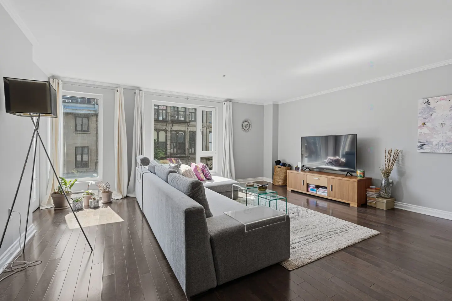 Modern living room with grey sofa, wooden TV stand, area rug, dark hardwood floors, and large windows with white curtains.
