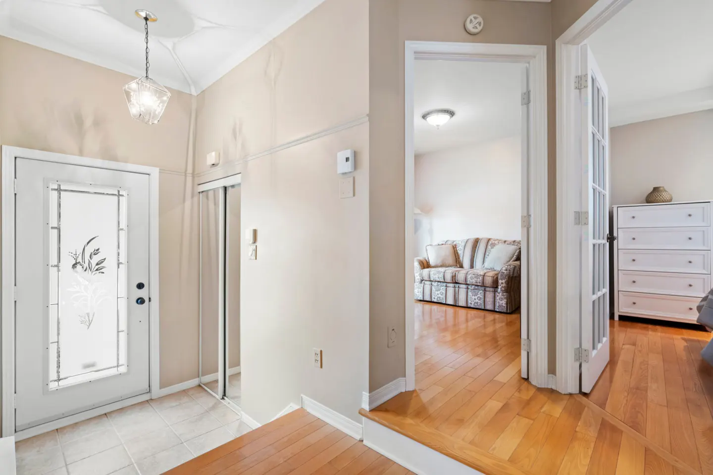 Foyer with a white front door with decorative glass, beige walls, and hardwood floors leading to a living room and bedroom.