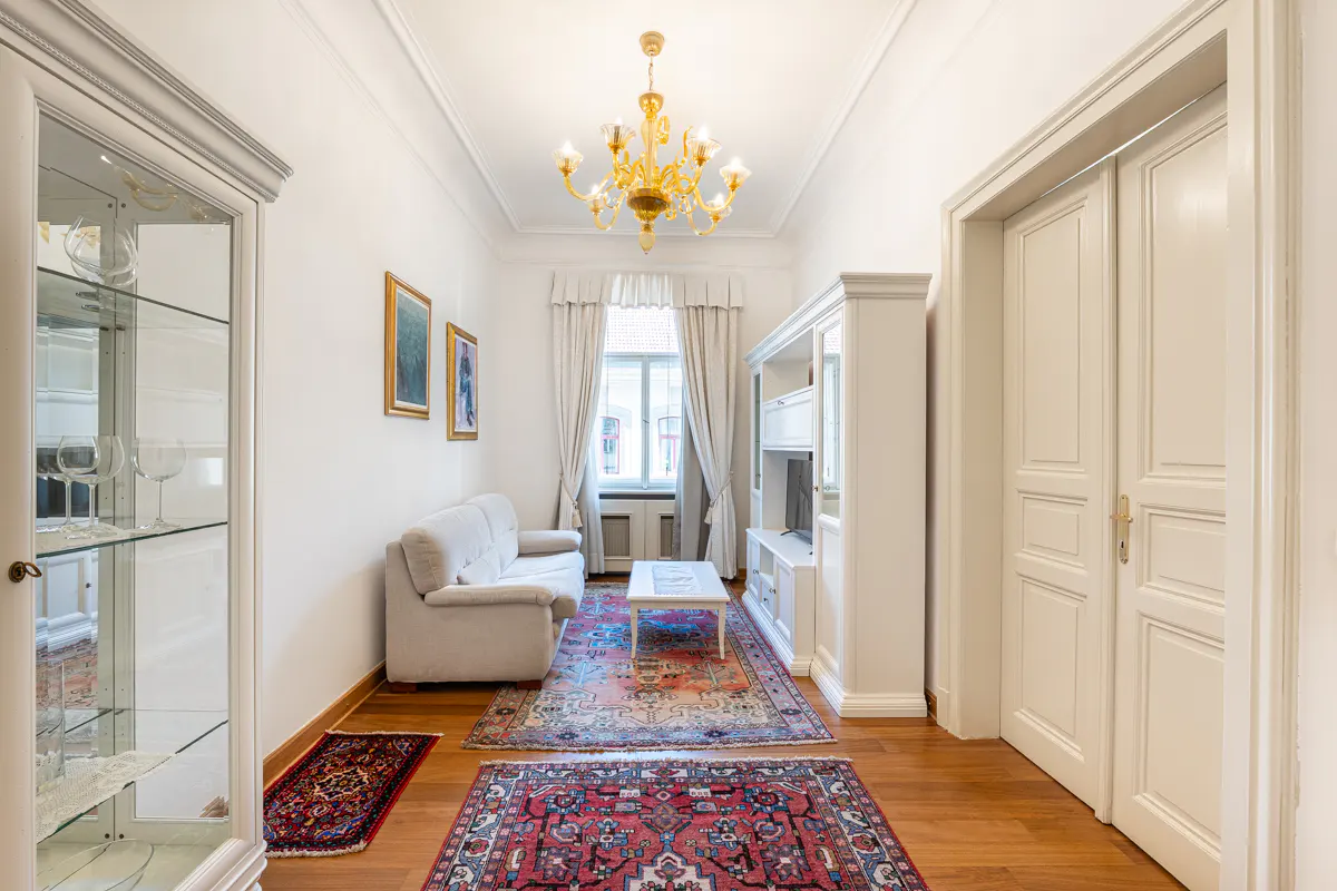 Bright living room with white walls, wood floors, and colorful rugs. A beige sofa sits near a window with white curtains. A gold chandelier hangs from the ceiling.
