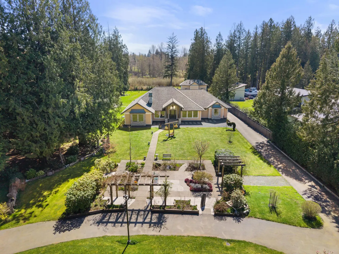 Aerial view of a one-story, light yellow house with a gray roof, surrounded by green lawns, trees, and a garden with pergolas.