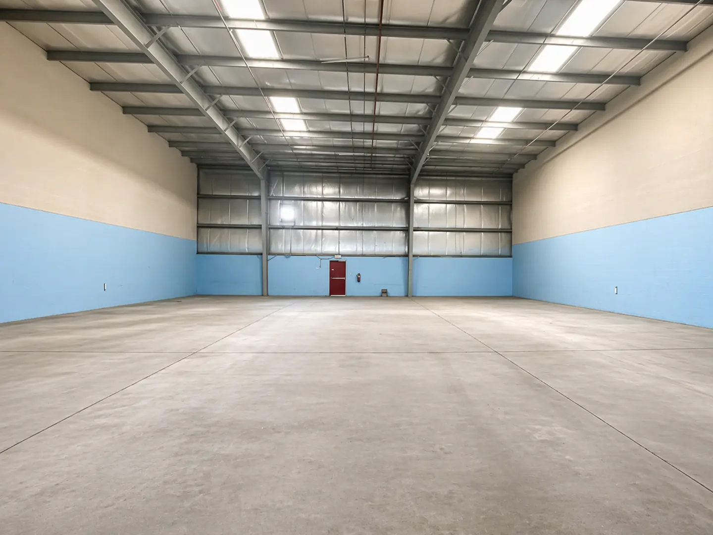 Empty warehouse interior with concrete floor, metal ceiling, and blue and beige walls. A red door is visible at the back.