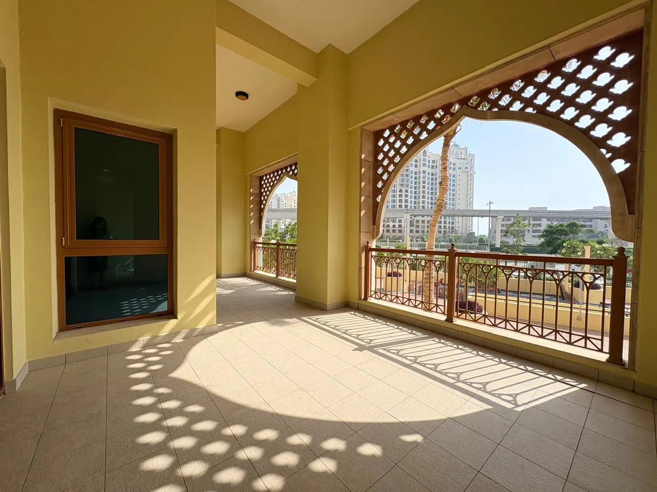 A sunny balcony with yellow walls, brown window frame, and decorative arched openings with city views.