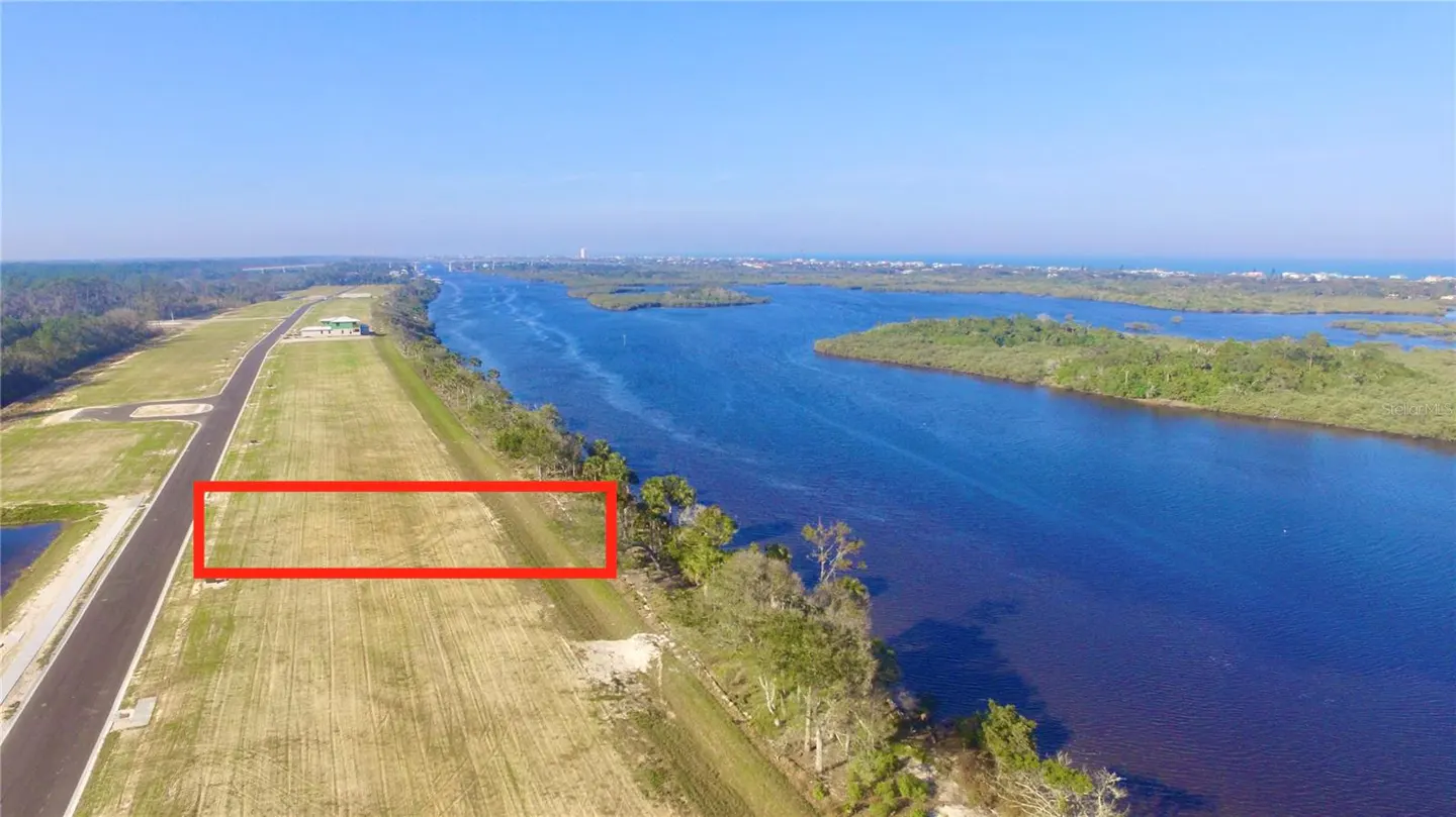 Aerial view of a rectangular grassy lot outlined in red, next to a blue river and green trees under a clear blue sky.