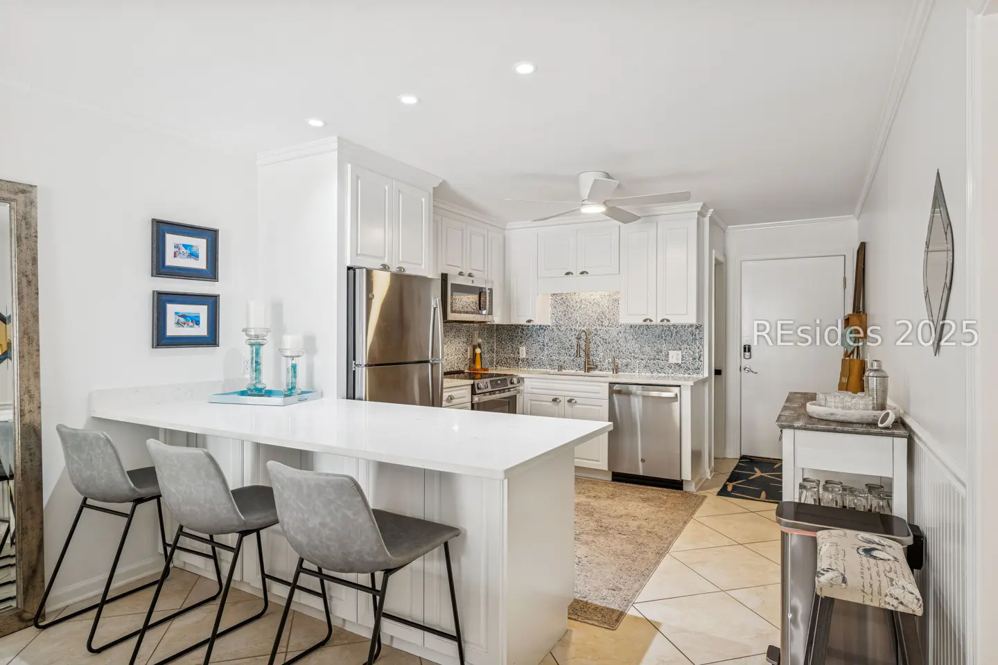 A bright kitchen with white cabinets, stainless steel appliances, and a white countertop island with three gray chairs.