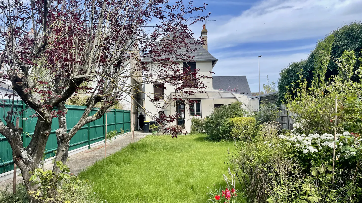 Exterior view of a two-story white house with a gray roof, green lawn, and lush garden. A tree with red leaves is in the foreground.