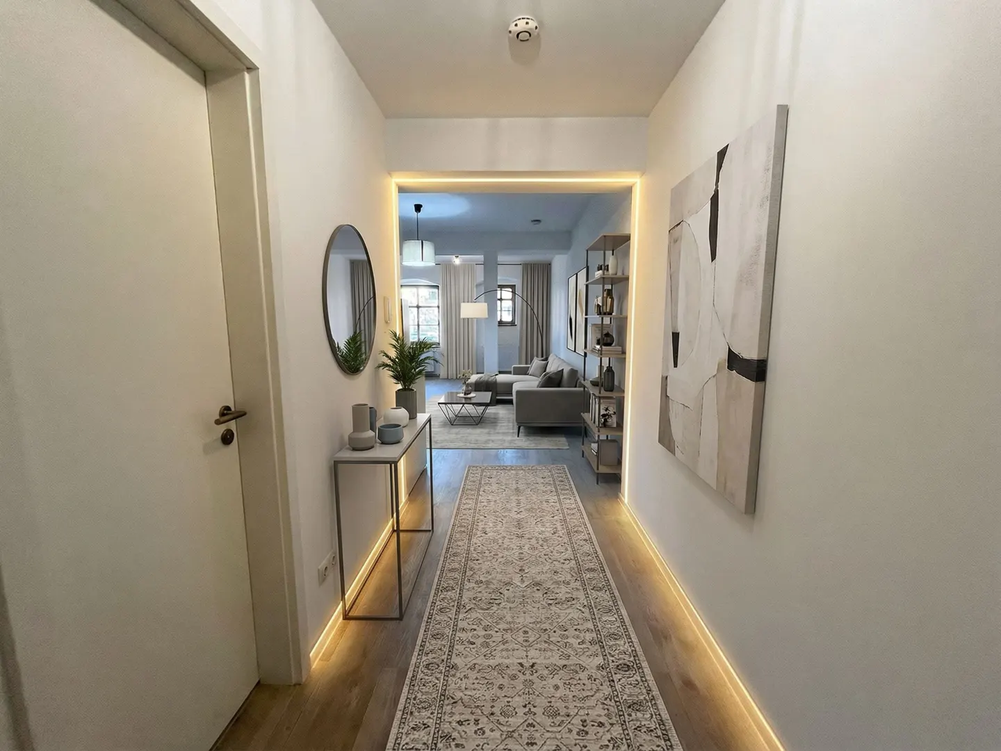 Hallway with a beige rug, white walls, and warm lighting. A console table with decor and a round mirror are on the left. An abstract painting is on the right. The living room is visible at the end of the hall.