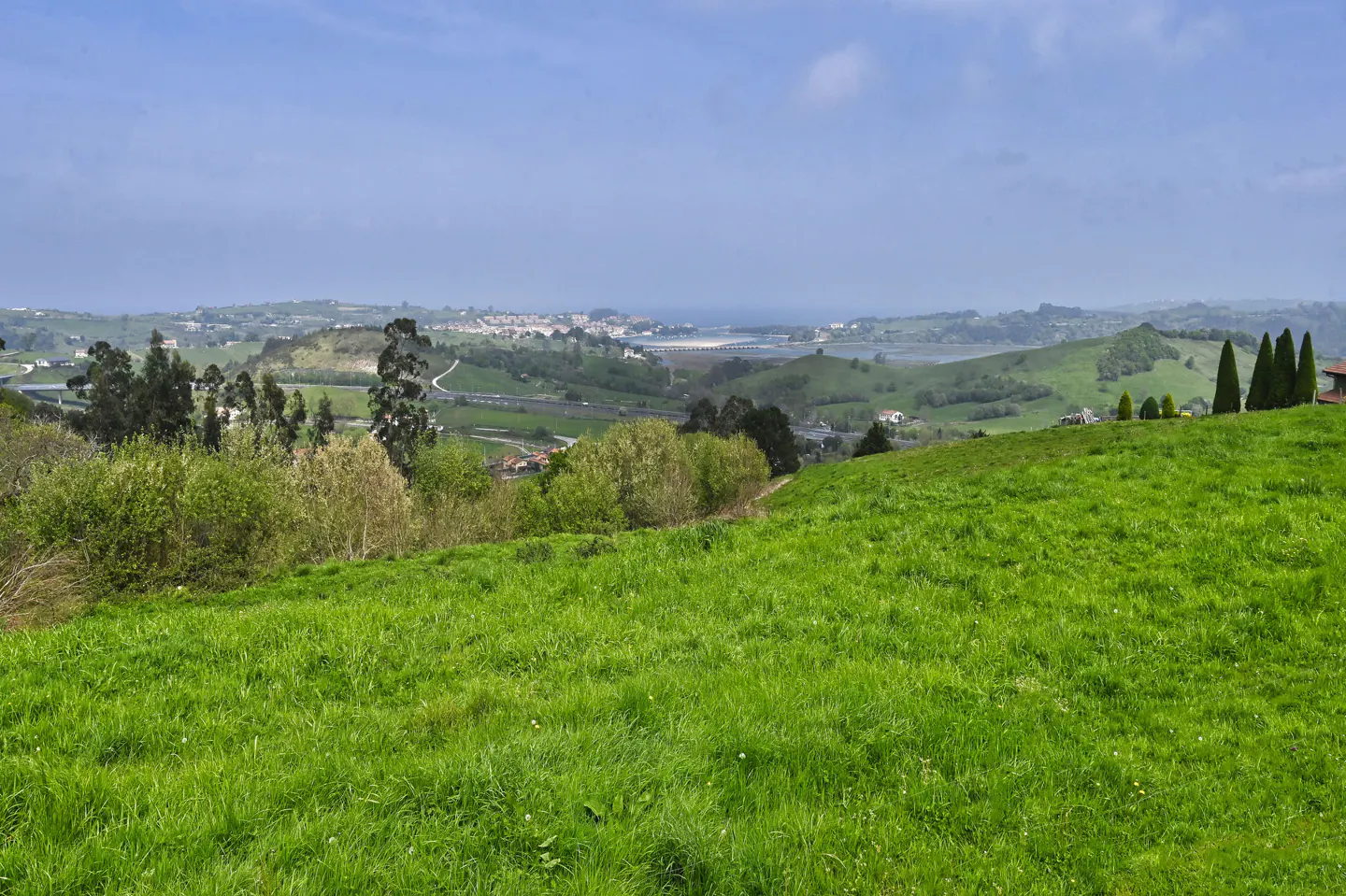 Scenic view of a green hillside overlooking a valley with trees, houses, and a distant town under a blue sky.