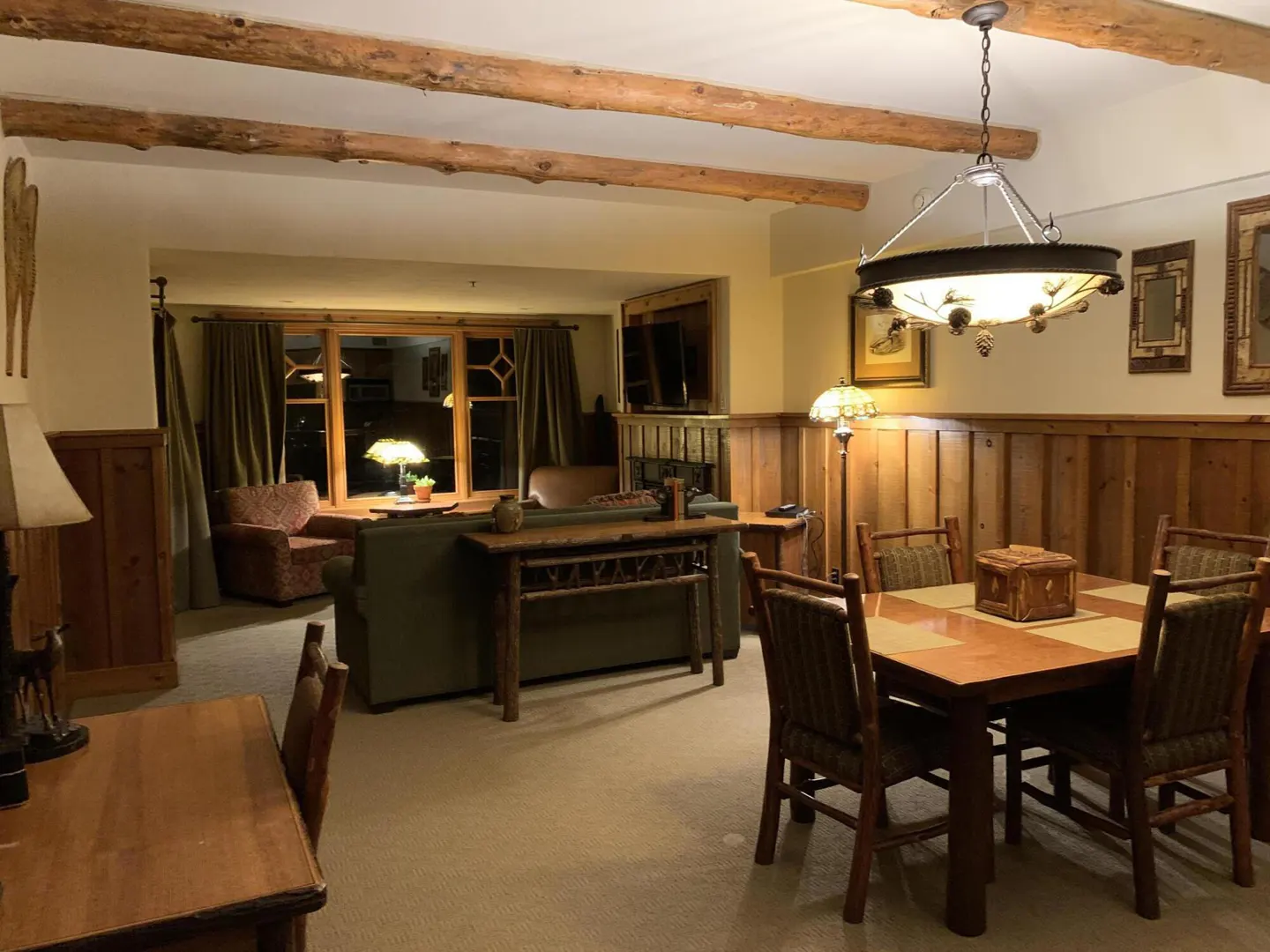 Living and dining area with wood paneling, exposed beams, and rustic furniture. A large window overlooks the night.