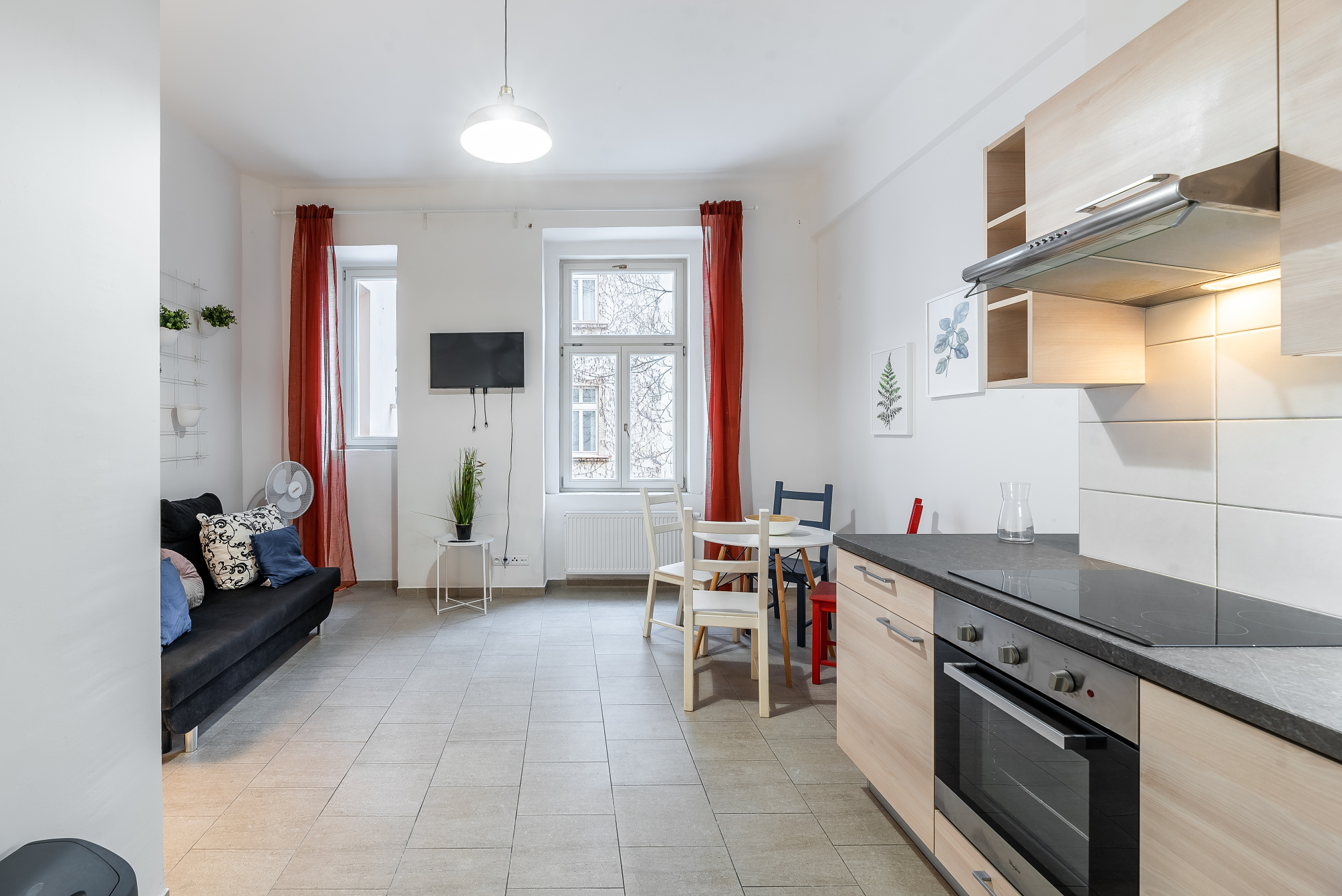 Modern studio apartment with a black sofa, dining area, and kitchen featuring light wood cabinets and a black stovetop. [AI-generated text]
