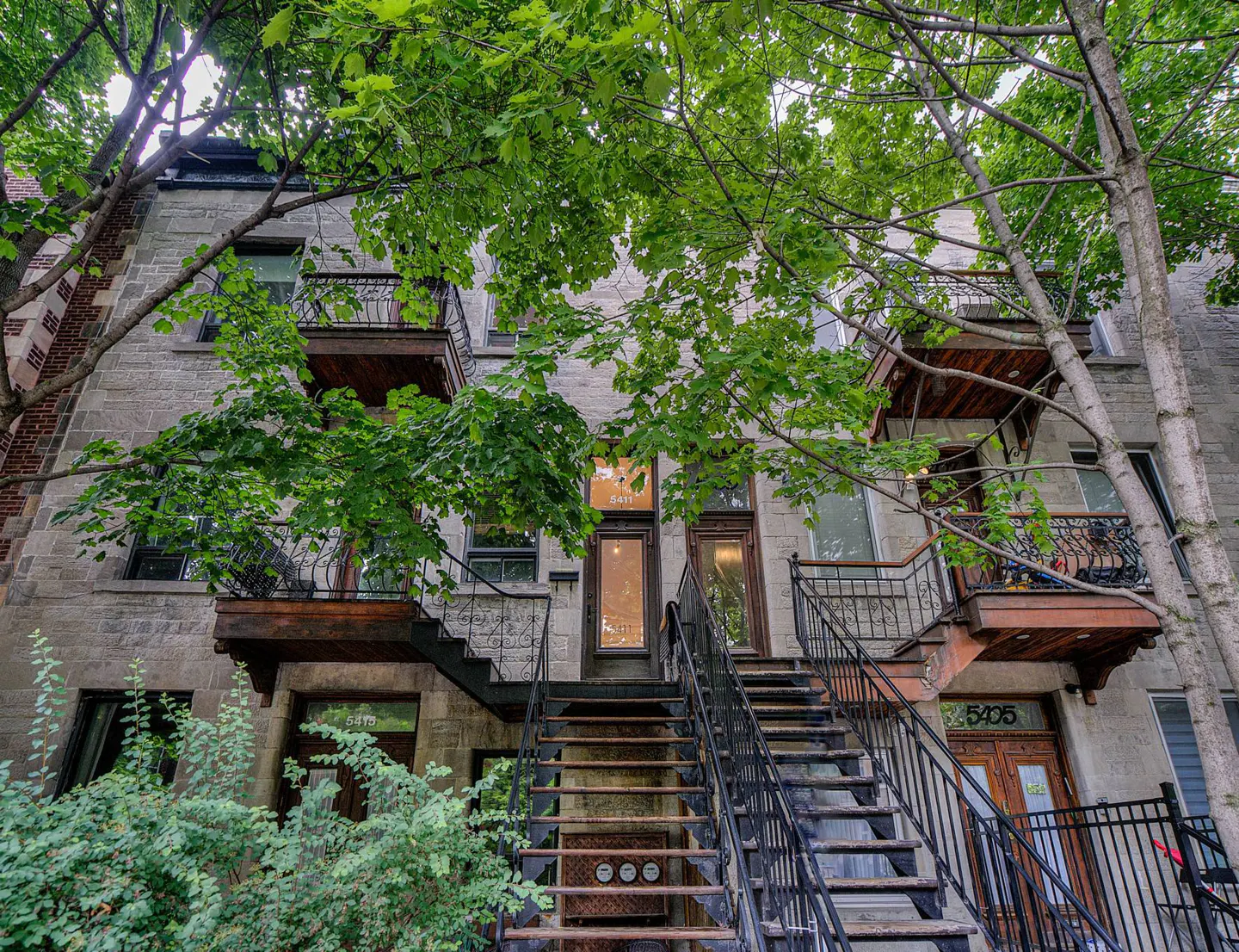 Exterior view of a three-story stone building with black iron staircases leading to each unit's entrance. Green trees frame the building.
