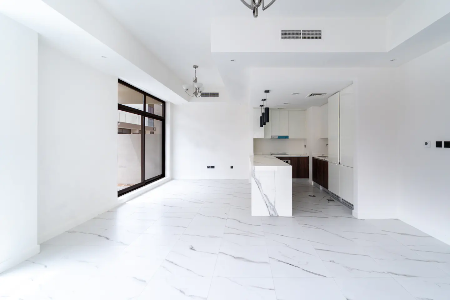 Bright, empty room with white walls, marble floors, and a kitchen with a marble island. A large window lets in natural light.