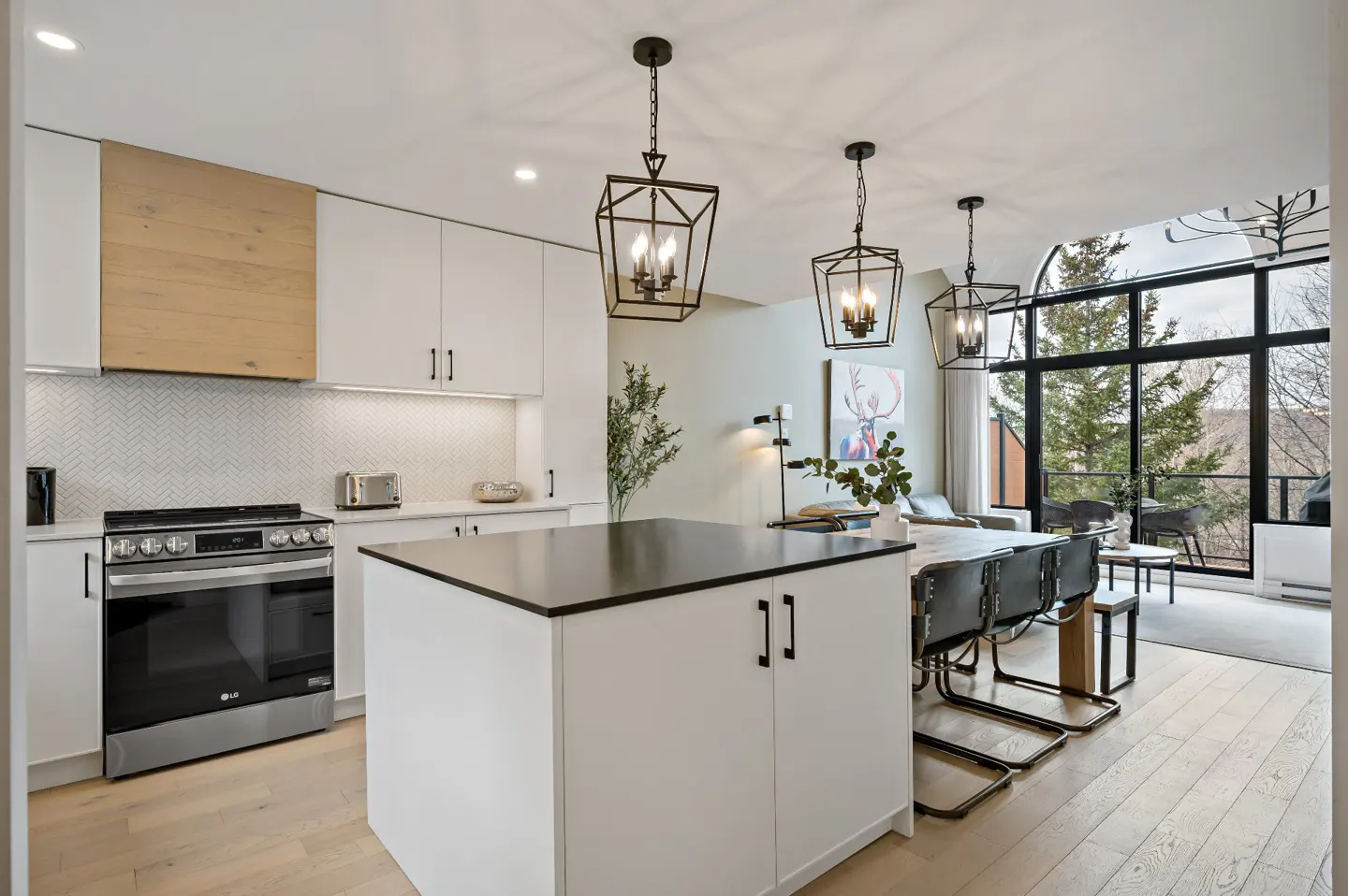 Bright, modern kitchen with white cabinets, black countertops, and stainless steel appliances. Three pendant lights hang above a central island. Large windows offer a view of trees.