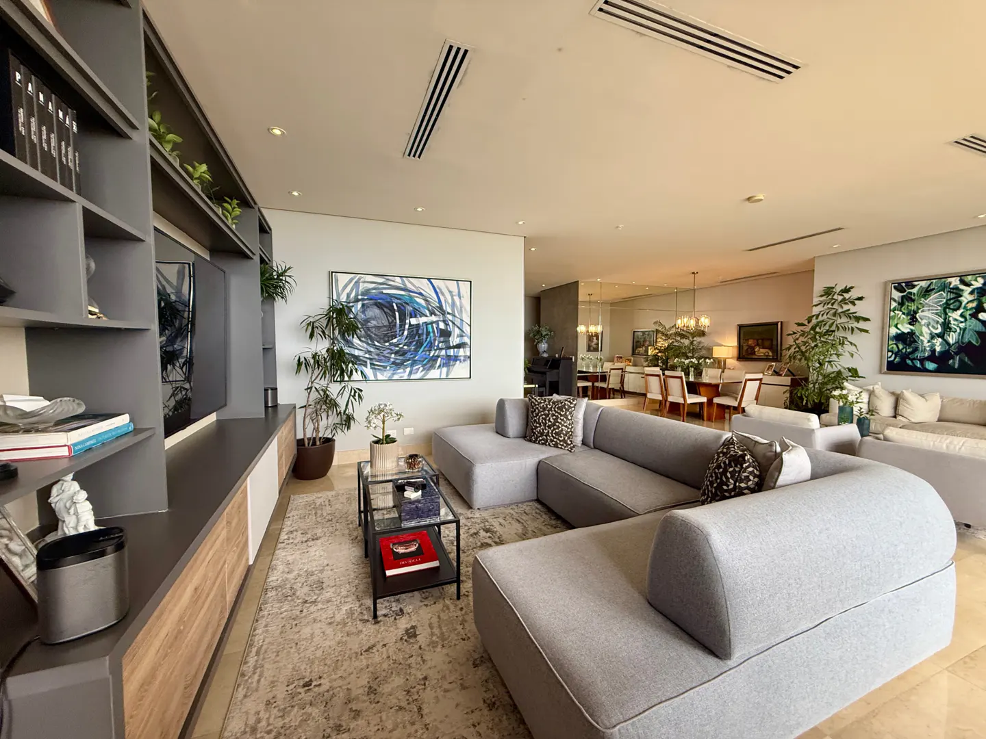 Bright living room with a gray sectional sofa, a glass coffee table, and a gray shelving unit with a TV. A dining area is visible in the background.