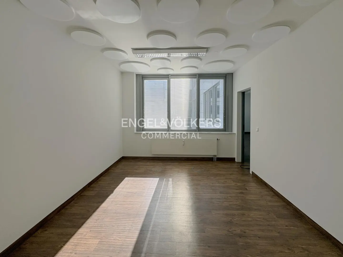 Empty office space with white walls, wood floor, and round ceiling lights. Windows let in sunlight. Engel & Völkers Commercial logo visible.