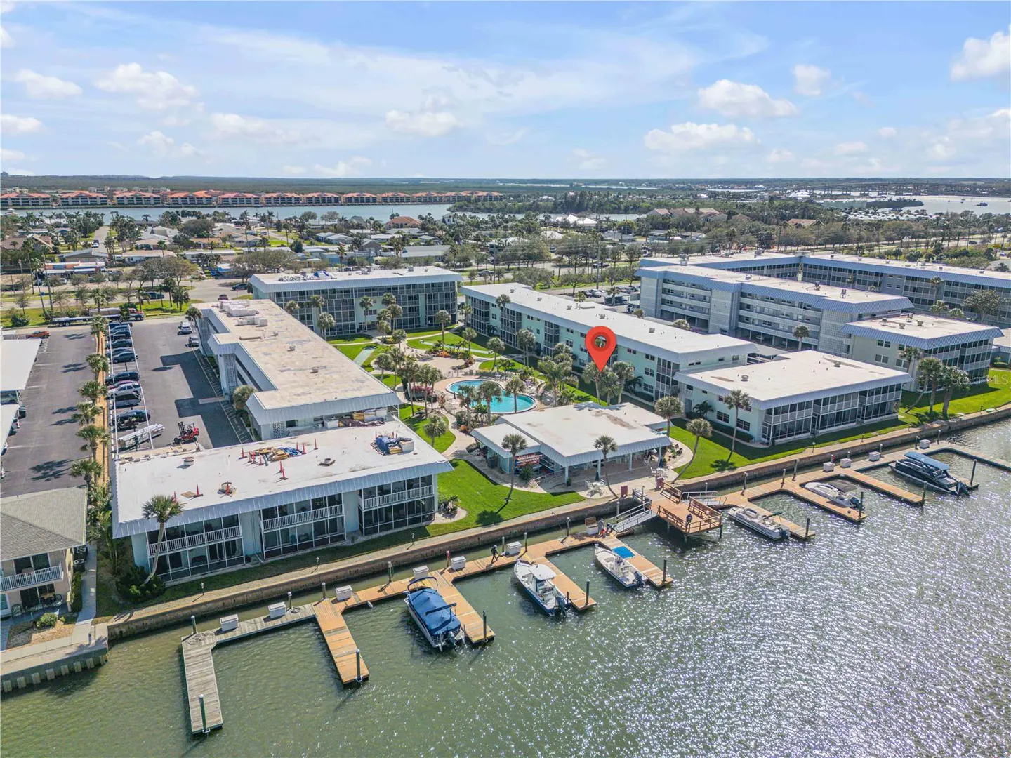 Aerial view of a waterfront condo complex with boat docks, a pool, and a parking lot under a blue sky.