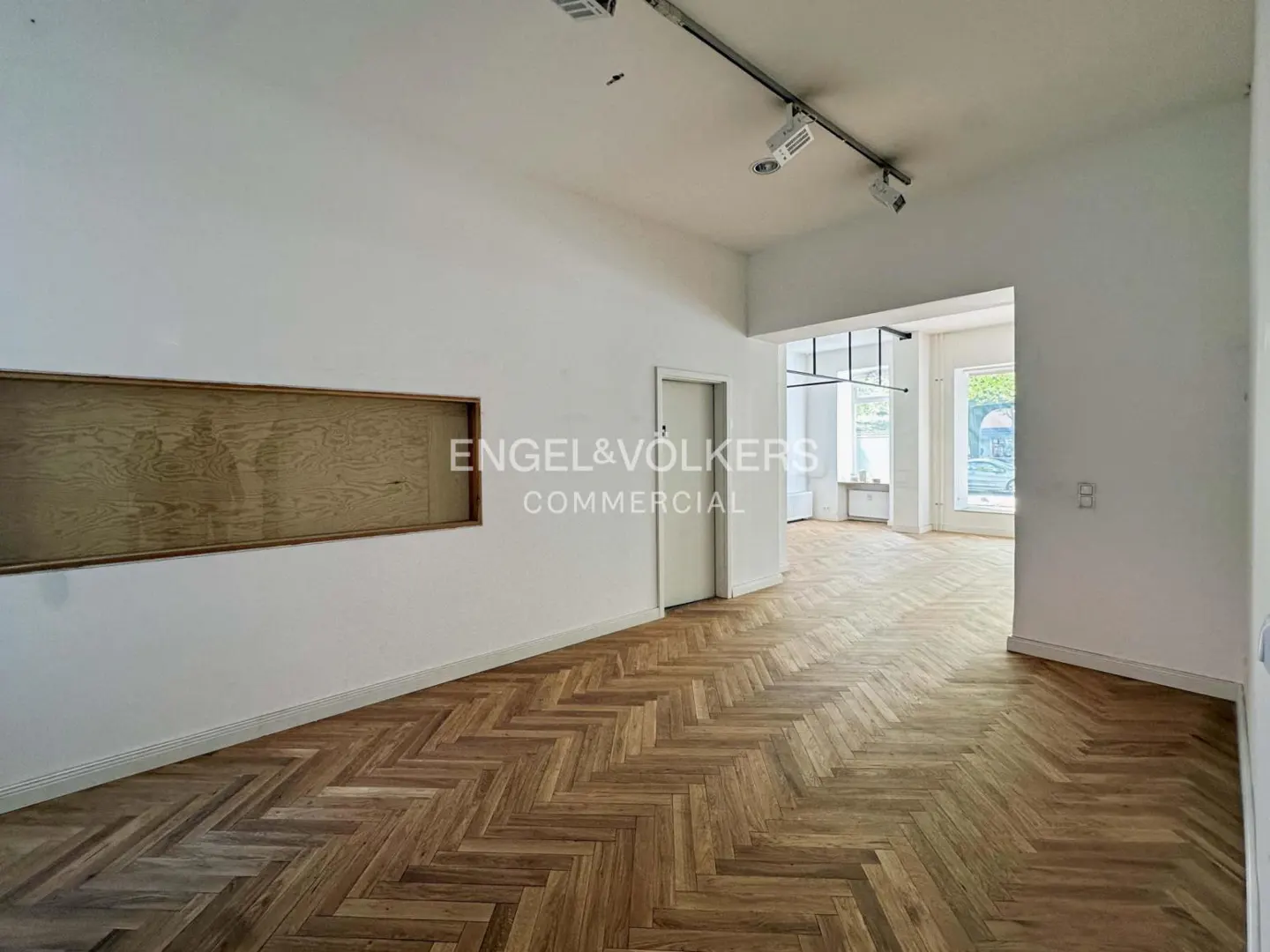 Empty commercial space with herringbone wood floors, white walls, and track lighting. Engel & Völkers logo on the wall.