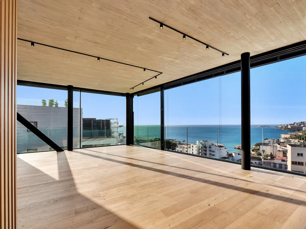 Bright, empty room with wood floors and ceiling. Black framed windows offer a panoramic view of the ocean and city. Track lighting on the ceiling.