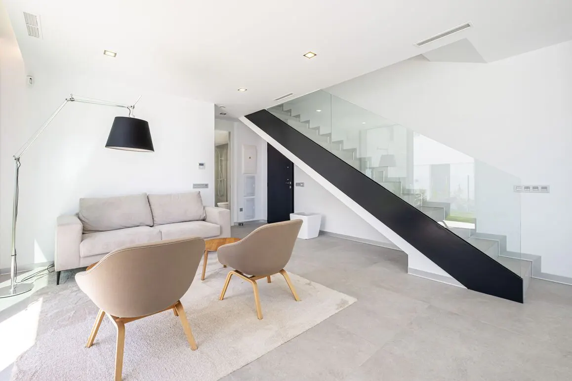 Bright living room with a beige sofa, two chairs, and a black and glass staircase.